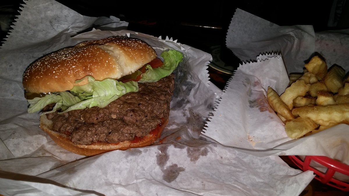 A cheeseburger with lettuce, tomato and pickles on a sesame seed bun, next to a basket of crinkle cut fries.