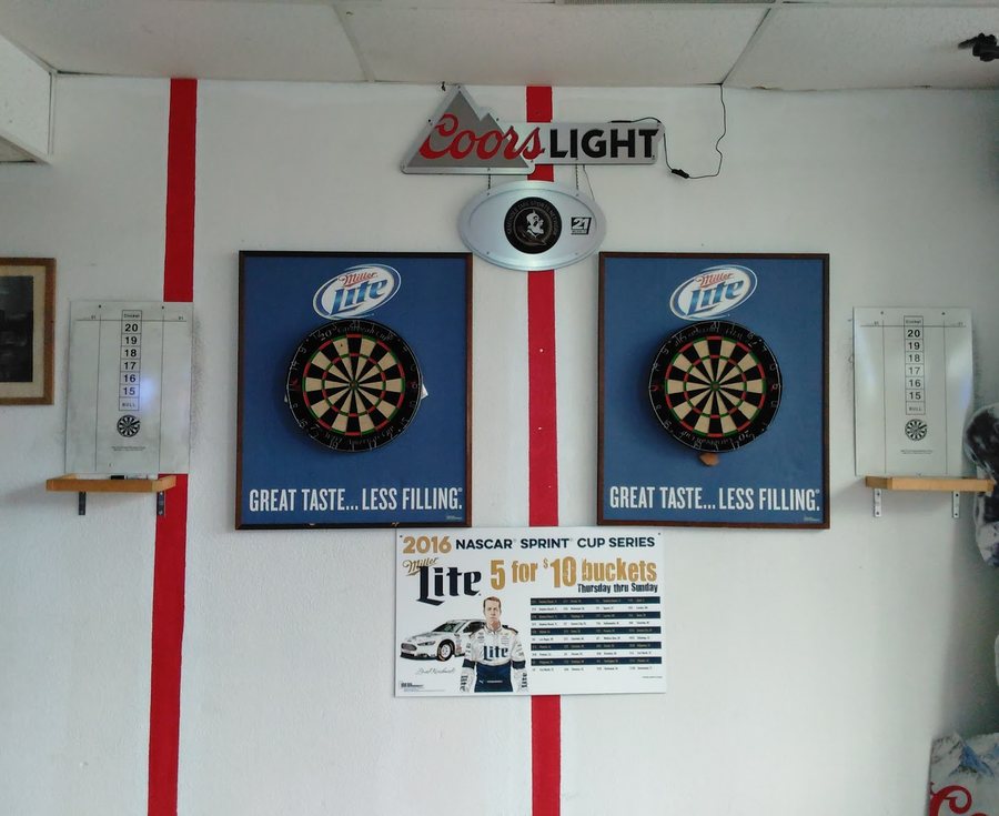 Two dartboards mounted on blue backboards under a lit Coors Light sign, with a paper scoreboard hanging on each side.