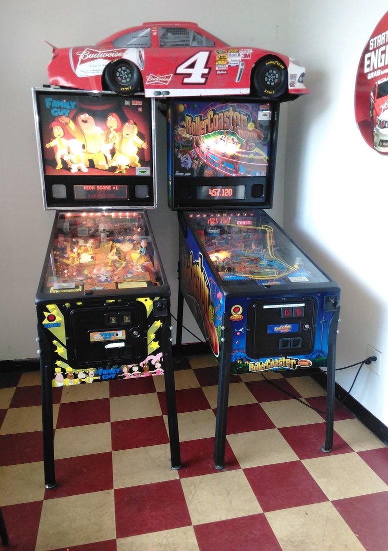 Two lit pinball machines side by side with a model stock car sitting on top of them, on the checkerboard floor.