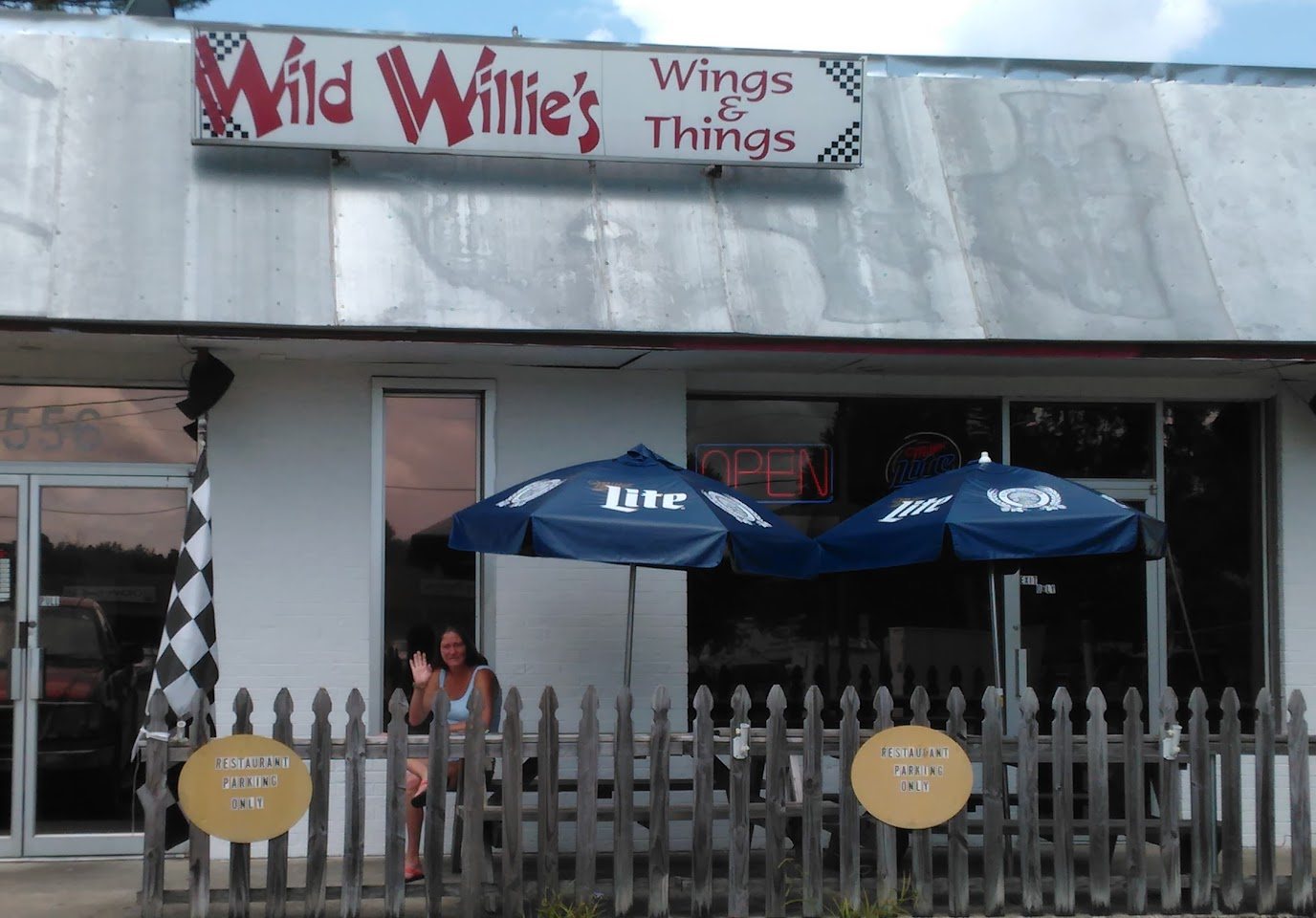 The front of the building: a red and white Wild Willie’s Wings & Things sign over a white storefront, blue umbrellas and a picket fence by the door, and someone waving from the window.