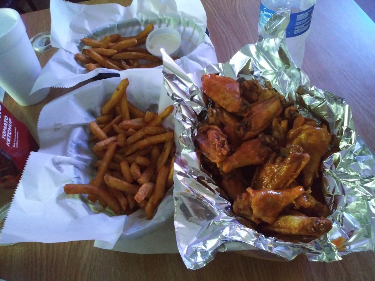 A foil basket heaped with sauced chicken wings beside two paper baskets of seasoned fries and a cup of dipping sauce.